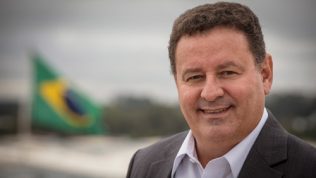 Um homem branco usando terno e camisa social sorrindo olhando fixamente para a câmera em fundo desfocado