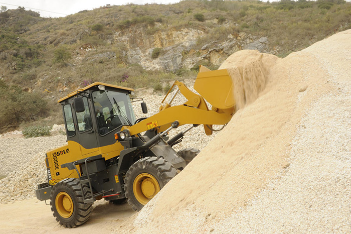 Cargador SDLG LG918, arena desbordante, con brazo de máquina a media altura. Al fondo, vegetación baja sobre un cerro.