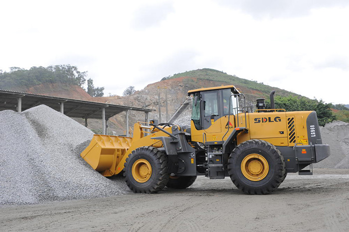 Cargadora de ruedas SDLG LG968, recogiendo piedras, vegetación baja y algunas rocas en una colina al fondo.