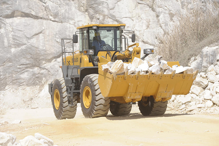 Cargadora de ruedas SDLG, transportando piedra, muro de piedra al fondo.