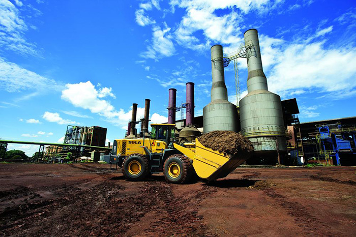 Cargador SDLG LG959, lleno de tierra, con una industria al fondo.