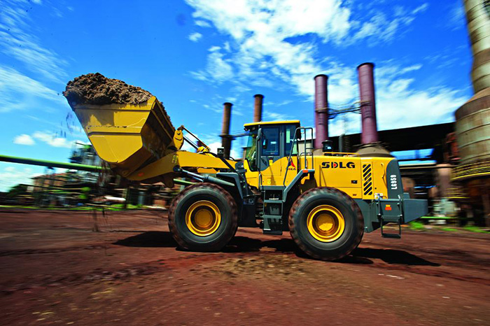 Cargador SDLG LG959, lleno de tierra, brazo de máquina levantado, con una industria al fondo.