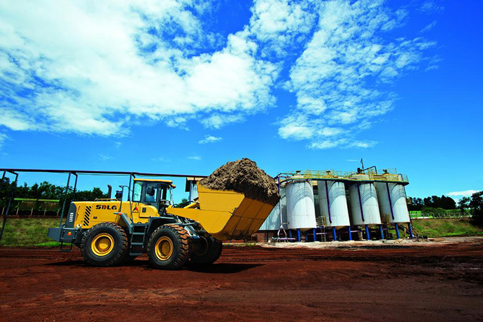 Cargador SDLG LG959, lleno de tierra, con una industria al fondo y un cielo azul.