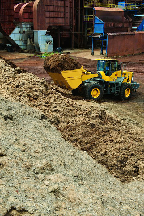 Cargadora de ruedas SDLG LG959, transportando tierra, con la industria al fondo.