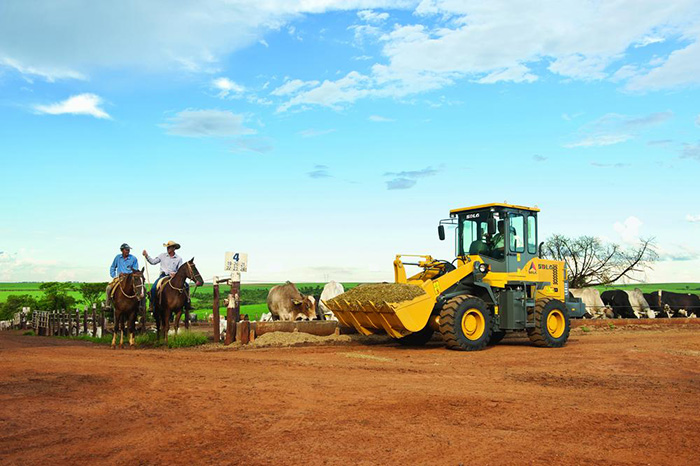 Cargadora SDLG LG918, transportando alimentos para el ganado al borde de una cerca. Al costado, dos hombres montados a caballo y al fondo, pasto con un cielo azul.