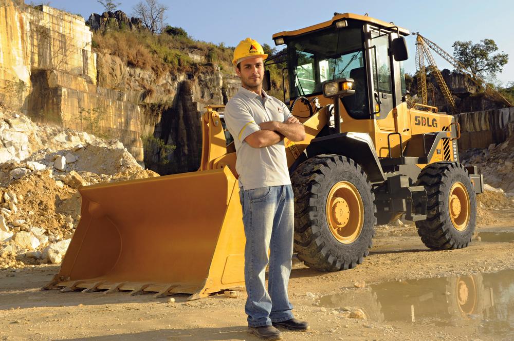 Un hombre blanco con una camiseta, jeans y un casco amarillo, en el fondo, cargadora de ruedas SDLG LG936L
