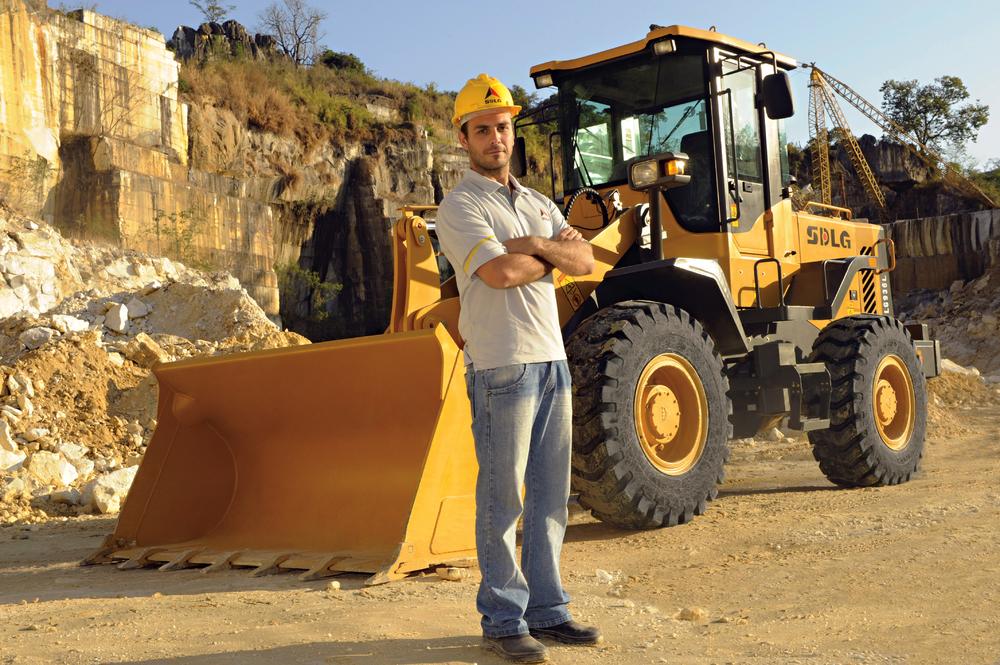 Un hombre blanco con una camiseta, jeans y un casco amarillo, en el fondo, cargadora de ruedas SDLG