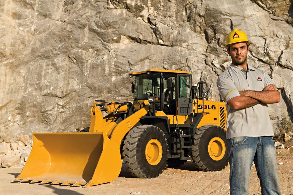 Un hombre blanco con una camiseta, jeans y un casco amarillo, en el fondo, cargadora de ruedas SDLG