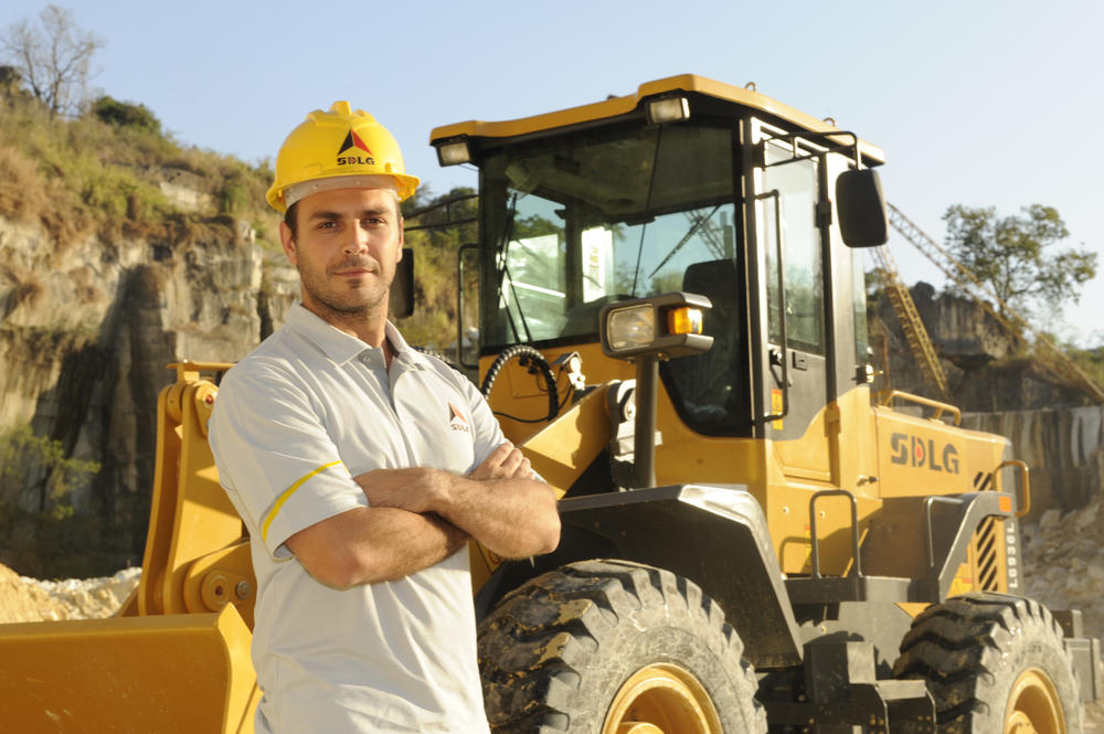 Un hombre blanco con una camiseta, jeans y un casco amarillo, en el fondo, cargadora de ruedas SDLG LG936L
