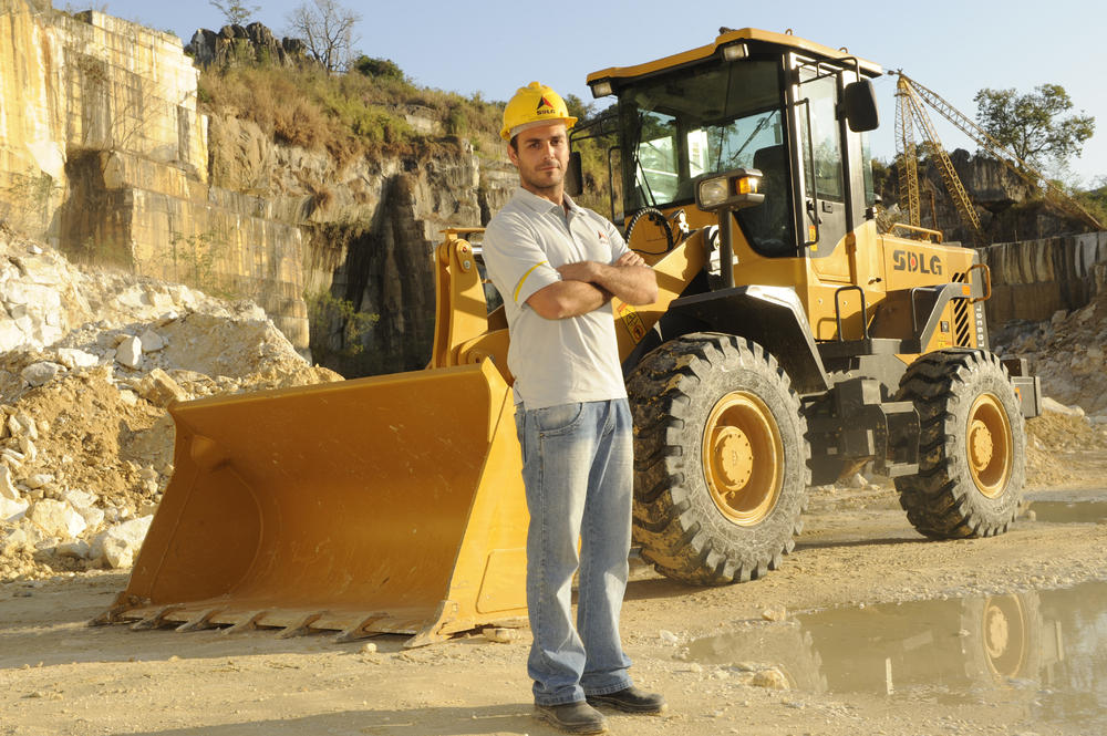 Un hombre blanco con una camiseta, jeans y un casco amarillo, en el fondo, cargadora de ruedas SDLG LG936L
