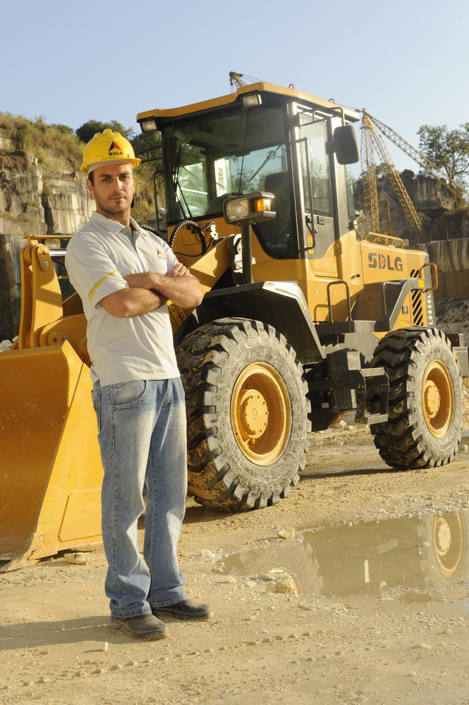Un hombre blanco con una camiseta, jeans y un casco amarillo, en el fondo, cargadora de ruedas SDLG LG936L