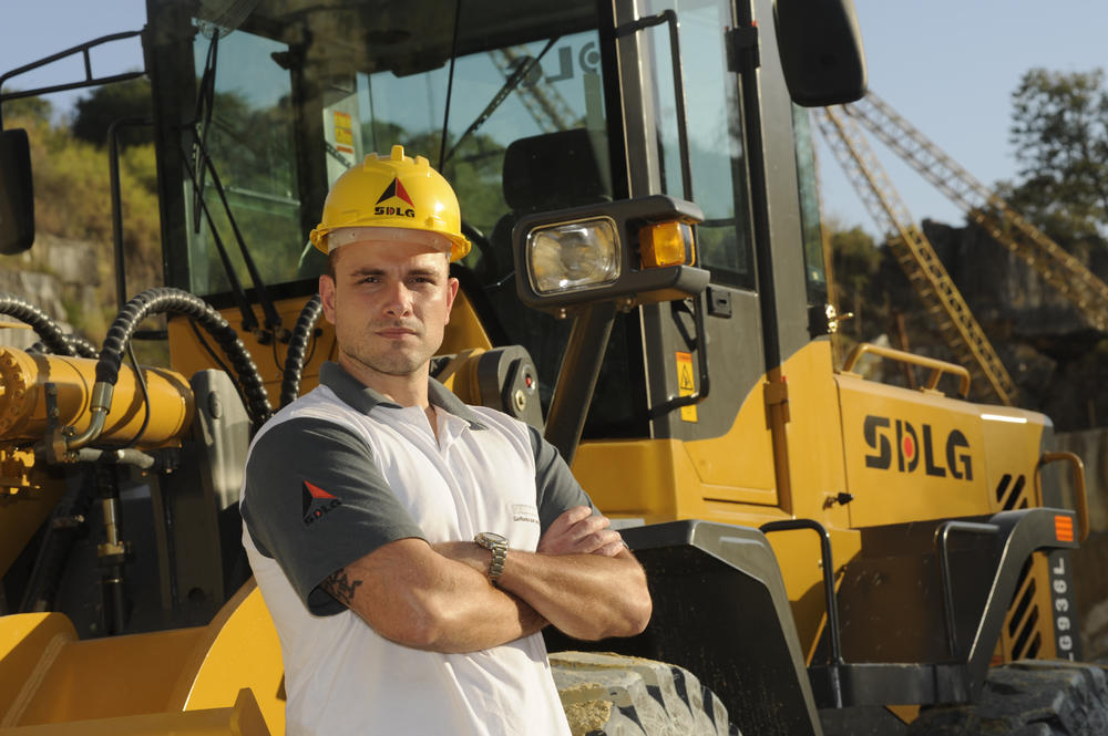 Un hombre blanco con una camiseta y un casco amarillo, en el fondo, cargadora de ruedas SDLG LG936L