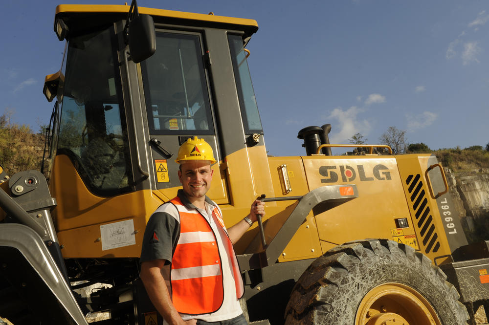 Un hombre blanco con una camiseta, un chaleco naranja y um casco amarillo sonriendo, de pie junto a uma cargadora SDLG 