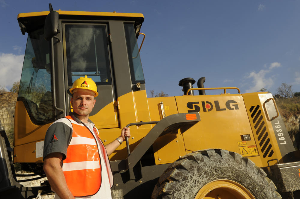 Un hombre blanco con una camiseta, un chaleco naranja y um casco amarillo, de pie junto a uma cargadora SDLG 