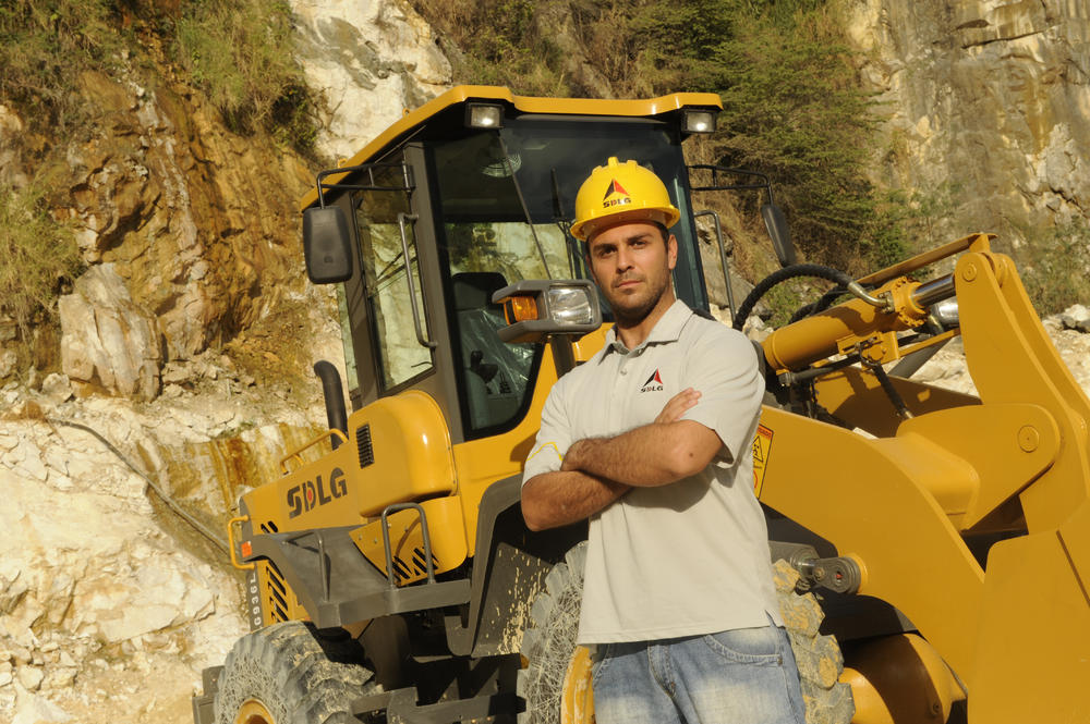 Un hombre blanco con una camiseta, jeans y un casaco amarillo se para junto a um cargador SDLG en el fondo de una montanã rocosa