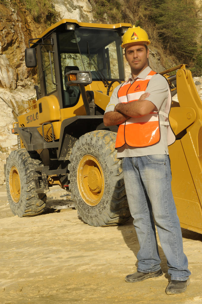 Un hombre blanco con una camiseta, jeans, un chaleco naranja y un casaco amarillo se para junto a um cargador SDLG en el fondo de una montanã rocosa