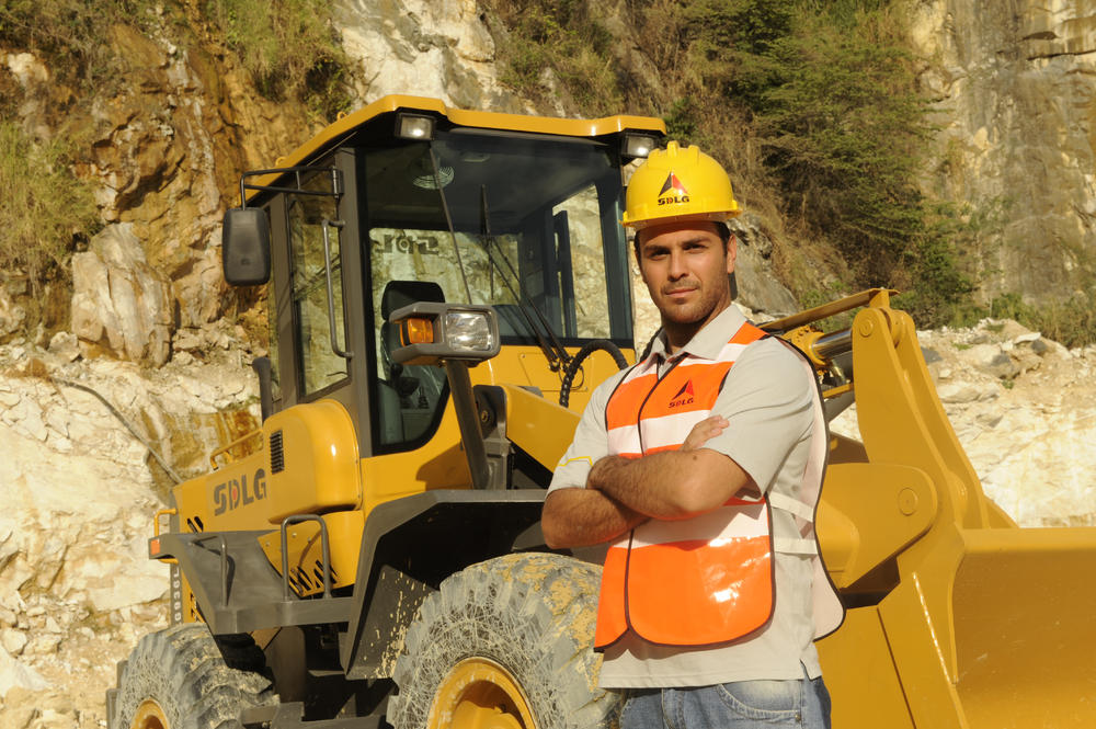 Un hombre blanco con una camiseta, jeans, un chaleco naranja y un casaco amarillo se para junto a um cargador SDLG en el fondo de una montanã rocosa