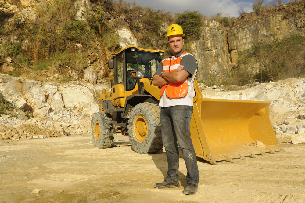 Un hombre blanco con una camiseta, jeans, un chaleco naranja y un casaco amarillo se para junto a um cargador SDLG en el fondo de una montanã rocosa
