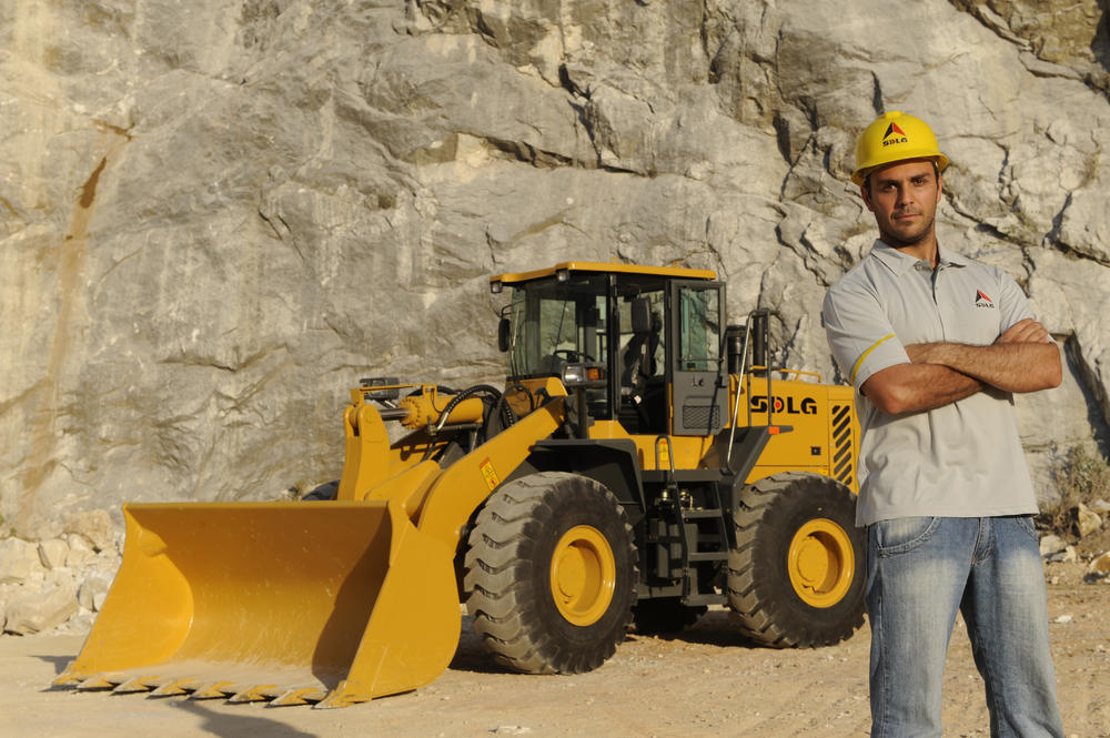 Un hombre blanco con camiseta, vaqueros y casco amarillo, ao fondo una cargadora SDLG y un muro de piedra