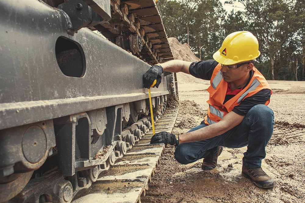Un trabajador verifica las medidas de la vía de la excavadora SDLG, árboles al fondo