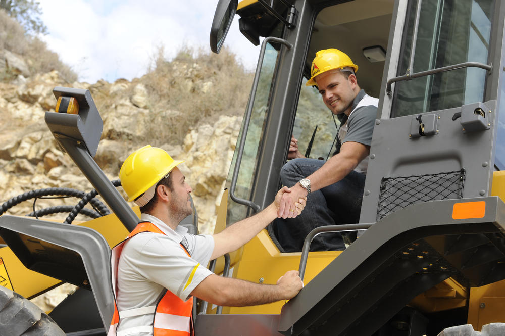 Un trabajador dentro del cargador SDLG le da mano a otro trabajador que está parado a un lado, al fondo piedras