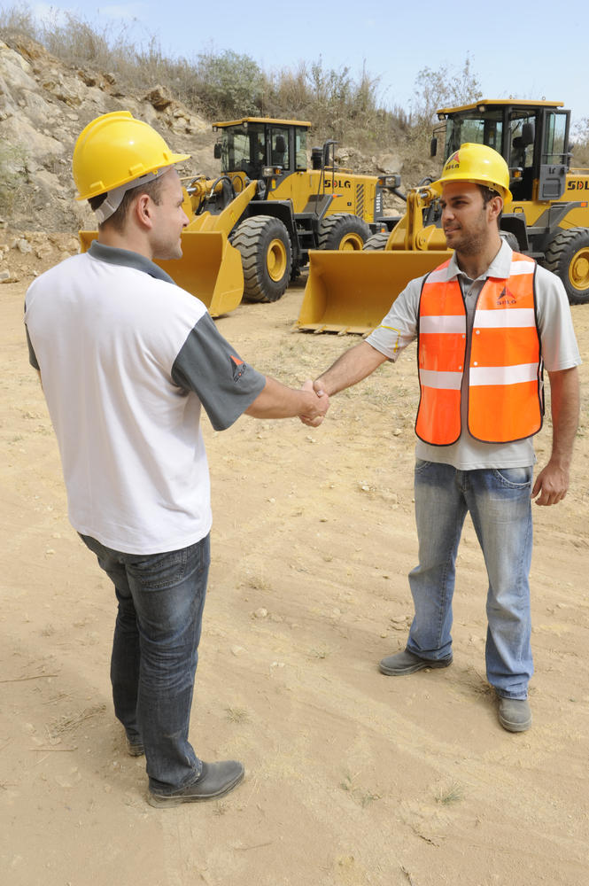Dos trabajadores uniformados y con cascos amarillos se dan la mano, al fondo cargadores SDLG y algunas piedras