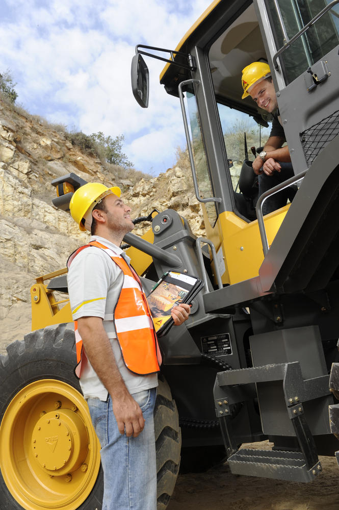 Un trabajador está dentro del cargador hablando con outro trabajador que está parado a un lado , al fondo un muro de piedra 