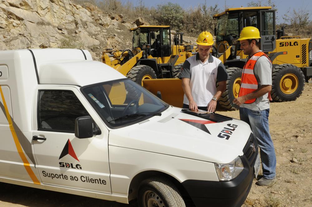 Dos trabajadores uniformados y con cascos amarillos hablan apoyados en el capó de un coche blanco con el logo de SDLG, al fondo cargadores de ruedas 
