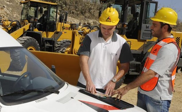 Dos trabajadores uniformados y con cascos amarillos hablan apoyados en el capó de un coche blanco con el logo de SDLG, al fondo cargadores de ruedas 