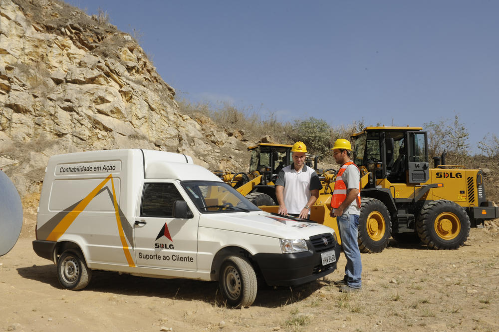 Dos trabajadores uniformados y con cascos amarillos hablan apoyados en el capó de un coche blanco con el logo de SDLG, al fondo cargadores de ruedas 