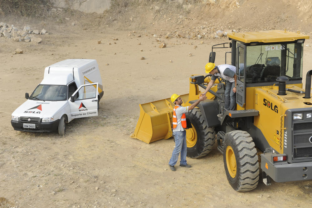 Dos trabajadores se saludan cerca de la cargadora SDLG, al fondo un coche blanco y um muro de piedra