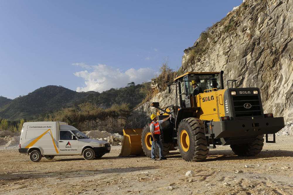 Dos trabajadores hablando, uno de ellos dentro de la percha del cargador y el otro de pie junto a un coche blanco y al fondo un muro de piedra