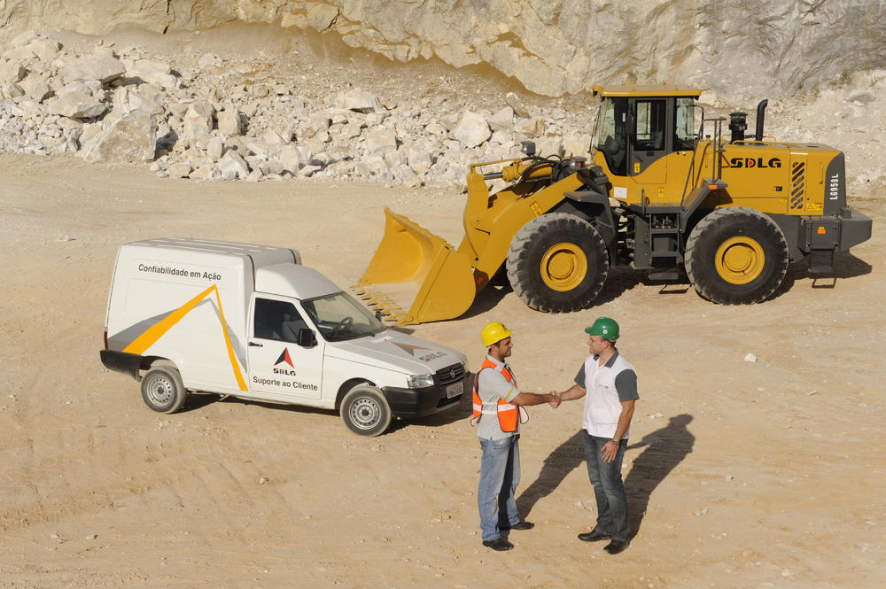 Dos trabajadores se dan la mano, al fondo un coche blanco, una cargadora SDLG y un muro de piedra