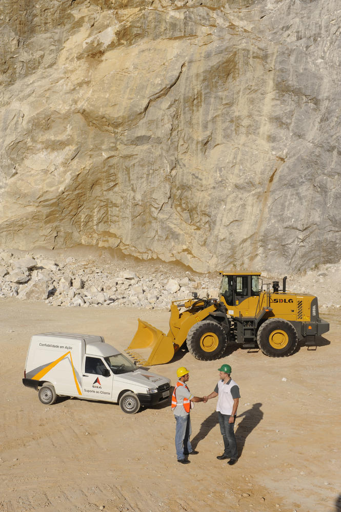 Dos trabajadores se dan la mano, al fondo un coche blanco, una cargadora SDLG y un muro de piedra