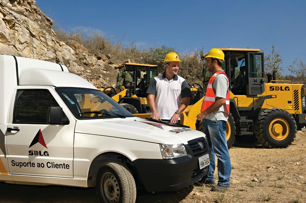 Dos trabajadores uniformados y con cascos amarillos hablan apoyados en el capó de un coche blanco con el logo de SDLG, al fondo cargadores de ruedas 