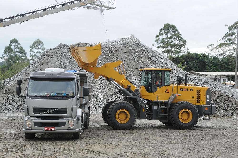 Cargadora de ruedas SDLG LG968 descargando rocas en un camión gris, con árboles y piedras en el fondo