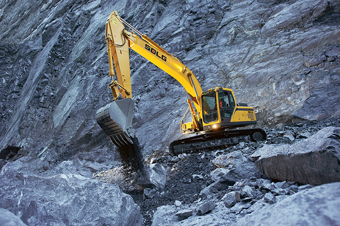 Excavadora SDLG, excavando tierra en un terreno irregular, con una pared de rocas al fondo.