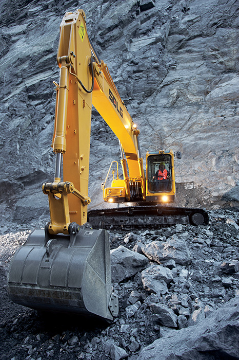 Excavadora SDLG, excavando tierra en un terreno irregular, con una pared de rocas al fondo.