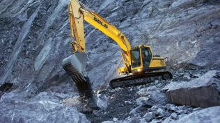 Excavadora SDLG, excavando tierra en un terreno irregular, con una pared de rocas al fondo.