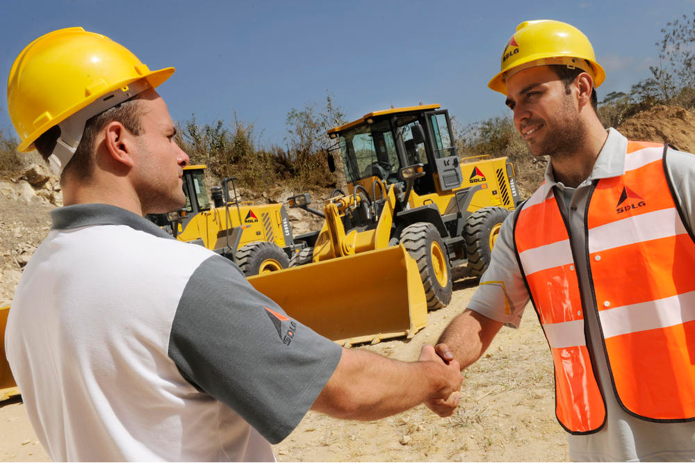 Dois homens brancos usando capacete amarelo se cumprimentam ao fundo escavadeiras SDLG