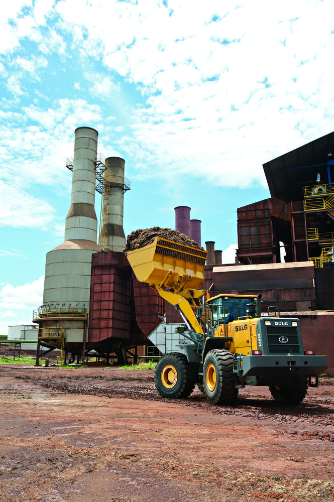 Cargador SDLG, lleno de tierra, con el brazo de la máquina levantado, al fondo una industria.