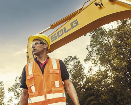 Um homem usando colete laranja e capacete amarelo olhando para algo, ao fundo parte do máquina SDLG