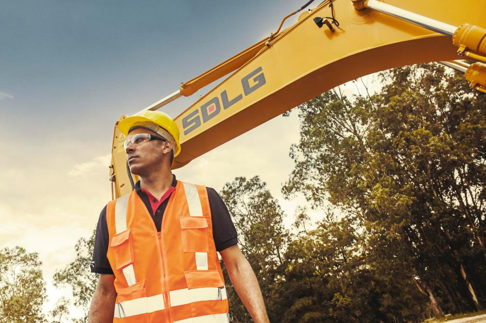 Um homem usando colete laranja e capacete amarelo olhando fixamente para algo ao fundo máquina SDLG