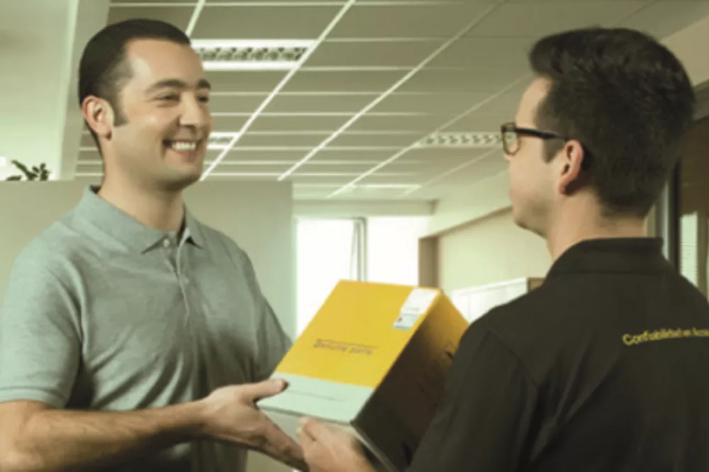 Dois homens brancos se cumprimentando sorrindo, segurando uma caixa amarela